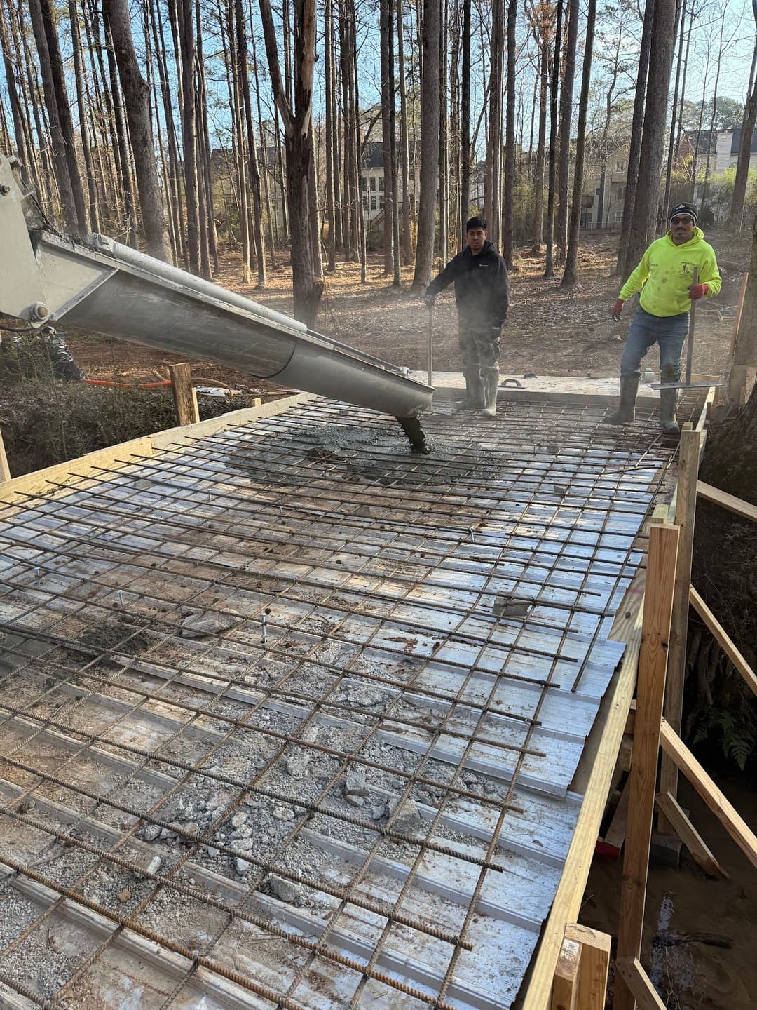 Workers pouring concrete onto a steel-reinforced slab in a wooded construction site.