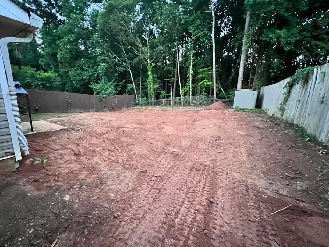 Cleared backyard area with dirt surface, surrounded by trees and a wooden fence.