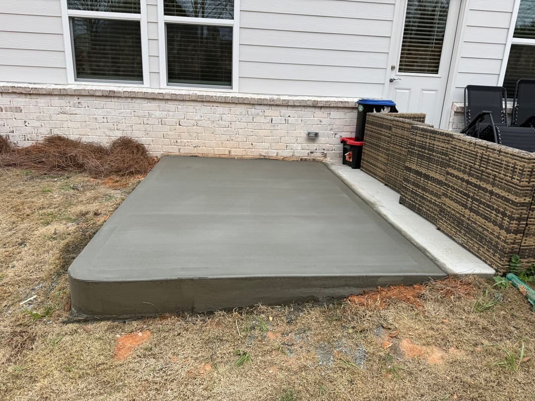 Freshly poured concrete patio slab beside a home, with grass and outdoor furniture visible.